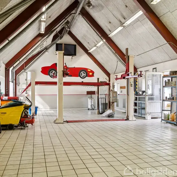 Redskaber og køretøjsløfter i aktivitet, placeret i en rummelig garage med skråt loft og flisegulv. Hylder med værktøj og rengøringsudstyr omkranser rummet. Ingen tekst fundet.