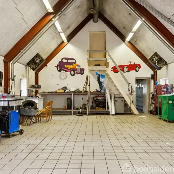 Et værksted med skrånende loft har farverige bilillustrationer på væggen. Midt i rummet er der en trætrappe op til en platform. Arbejdsborde og værktøj omkranser rummet.