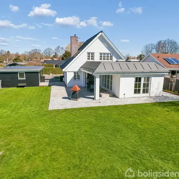 En hvid to-etagers villa med sort tag står midt i en velplejet grøn have. En lille terrasse med en udendørs pejs ses foran huset. Skyfri blå himmel.
