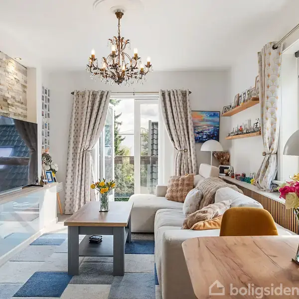 Stue med hvid sofa, sofabord og stor vægmonteret TV. Lyse gardiner pryder glasskydedøren til en balkon. Hylder, lamper og blomster udsmykker rummet. Chandelier hænger fra loftet.