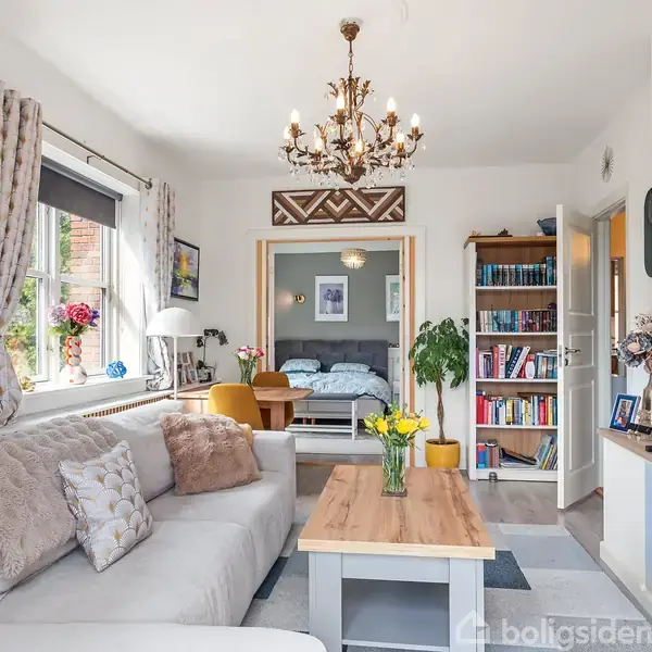 Stue med grå sofa og sofabord, pyntet med blomster. En lysekrone hænger over bordet. Der er en bogreol og indgang til et soveværelse bagved.