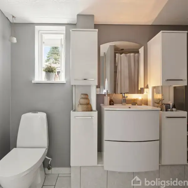 Toiletstol står stille i moderne badeværelse med grå vægge. Spejl og vask er flankeret af skabe. Et lille vindue lukker dagslys ind. Hvidt håndklæde hænger til højre.