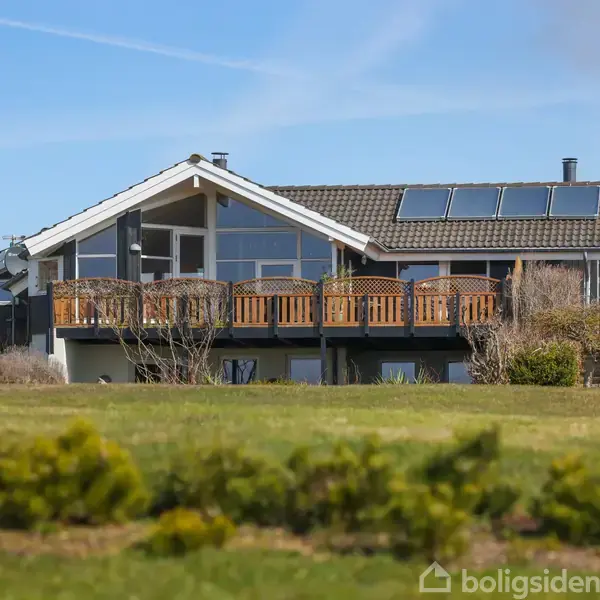 Et moderne enfamiliehus står på en velplejet græsplæne. Huset har en stor træterrasse og solpaneler på taget, omgivet af buske og træer under en klar blå himmel.