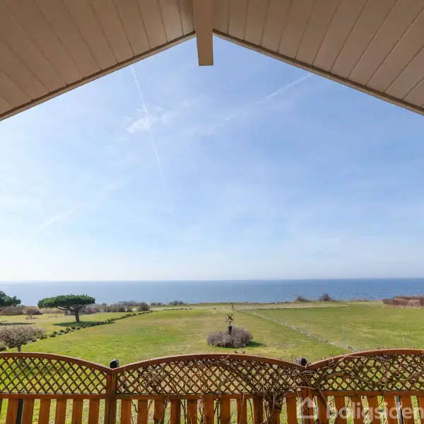 En veranda med trælignende gelænder giver udsigt over en stor, græsgrøn mark, der strækker sig til havet under en klar blå himmel.