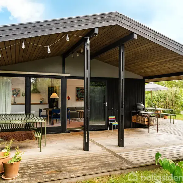 Sort træhus med overdækket terrasse, hvor et bord og stole står. Omgivet af grøn have med potteplanter. Loftet på terrassen er dekoreret med lyskæder. Ingen tekst i billedet.