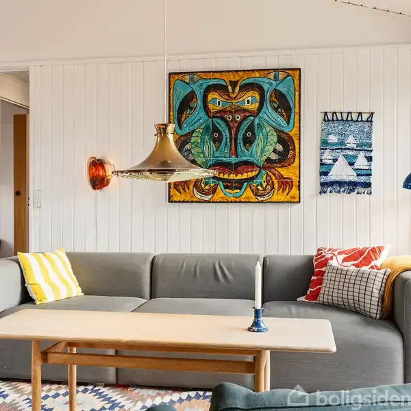 Gråt sofaarrangement med forskellige farverige puder står i stue. Over sofaen hænger en kunstnerisk plakat og lamper, mens et sofabord står foran. Panelvægge skaber en hyggelig atmosfære.