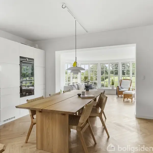 Spisebord med træstole står i et lyst køkken/alrum. En hængelampe hænger over bordet. I baggrunden ses et opholdsområde med store vinduer og udsyn til en have.