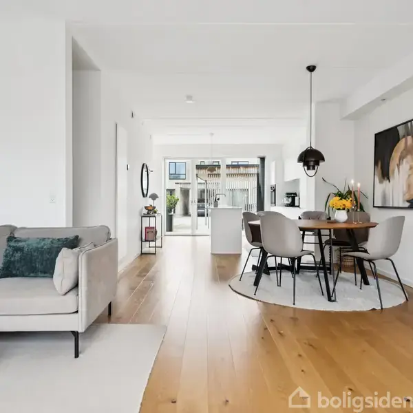 En lys stue med en grå sofa til venstre, et rundt spisebord med stole til højre, og et åbent køkkenområde i baggrunden. Moderne indretning med trægulv.