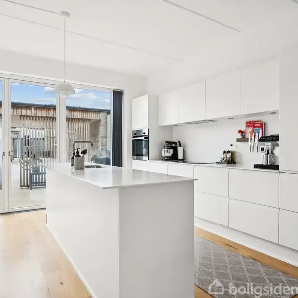 Hvidt køkken med ø, hvor kaffemaskine og diverse små apparater står på bordpladen. Store glasdøre åbner til en terrasse med planter og et hus i baggrunden.