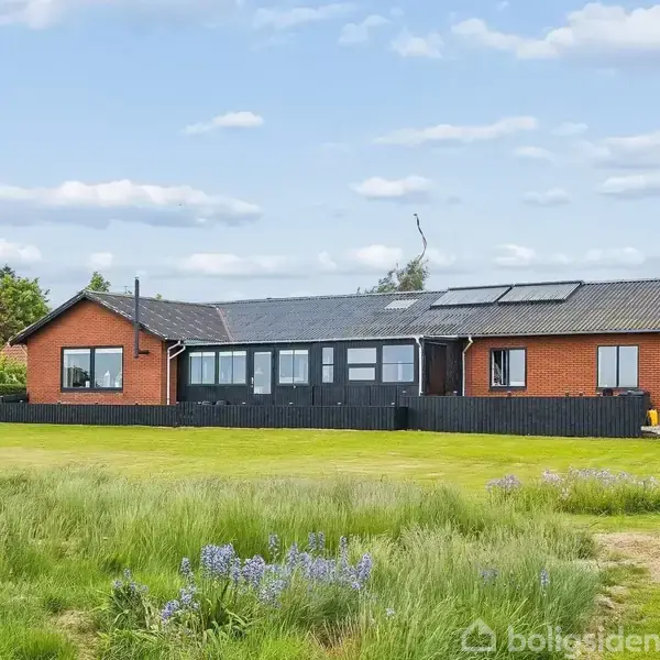 En rødstensvilla med solpaneler på taget står i en stor have med græsplæne og vilde blomster. Villaen har sorte rækværk og store vinduer. Himlen er let skyet.