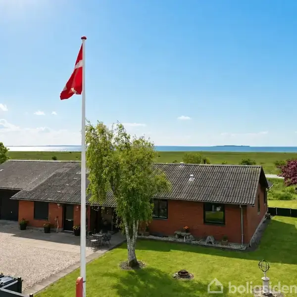 En rødstenshus med sorte tagsten står i en grøn have nær kysten. En dansk flagstang vajer stolt foran, omgivet af træer og blomster i et fredeligt landskab.