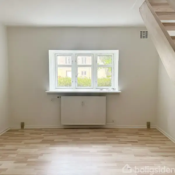 Et tomt rum med trægulv, hvor en trætrappe fører opad. Et vindue i baggrunden lader lys trænge ind. Radiator under vinduet, lyse vægge.