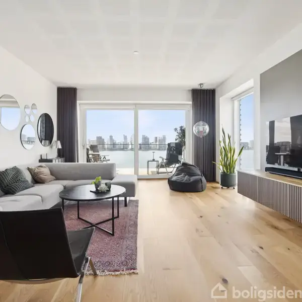 Stue med moderne indretning; en grå sofa og sort lænestol omkring et rundt bord, stor fladskærm på væggen; store vinduer med udsigt over byen; træplankegulv.