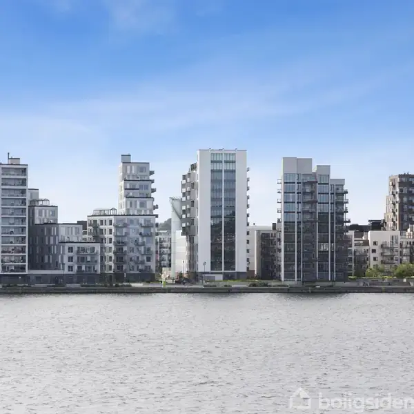 En række moderne lejlighedsbygninger står langs en flod. Bygningerne har forskellige højder og designs, som afspejler sig i vandet under en klar blå himmel.