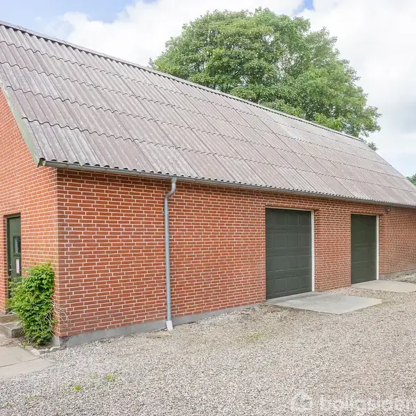 En murstensbygning med to garagedøre står stille i et grønt landskab. Stier omkranser bygningen, der har et skråt tag og grus på jorden foran.