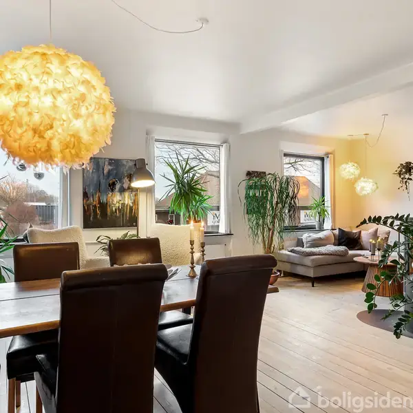 En spisestue med et bord omgivet af stole, oplyst af en stor gul lampe. Rummet er fyldt med grønne planter og har adgang til en stue med sofaer.