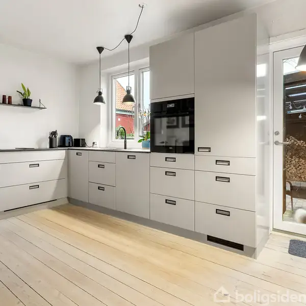 Køkken med lysegrå skabe og indbygget ovn. Tre hængende lamper lyser rummet op. Store vinduer giver udsigt til en terrasse med møbler. En hvid pelspude ligger på en taburet.