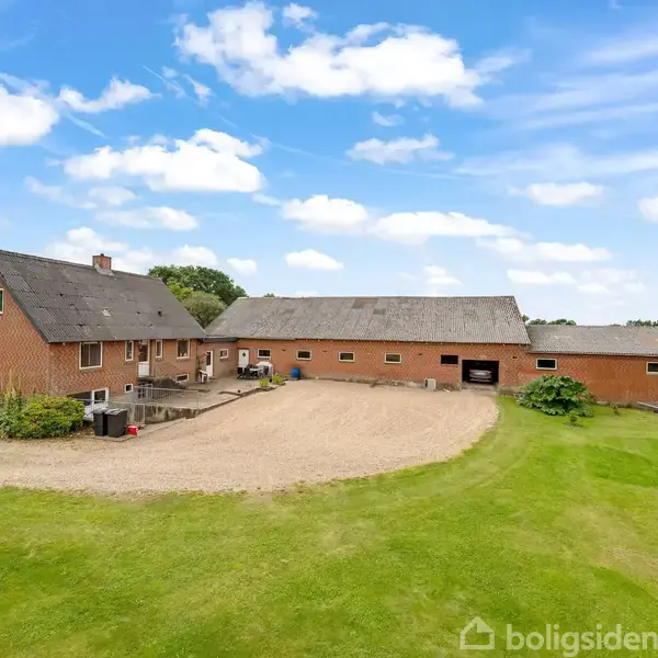 Murstenshus og lade står ved siden af hinanden, omgivet af en grusvej og grøn græsplæne, under en klar blå himmel med skyer.