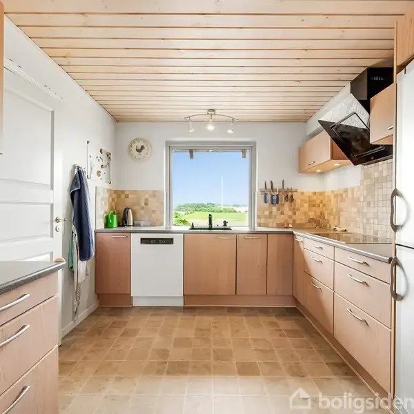 Køkken med træskabe og flisegulv, opvaskemaskine ved vinduet, der viser udsigt til græsmark. Vægge med lyse fliser, skabe med stålhåndtag, og åben glaslåge i skab.