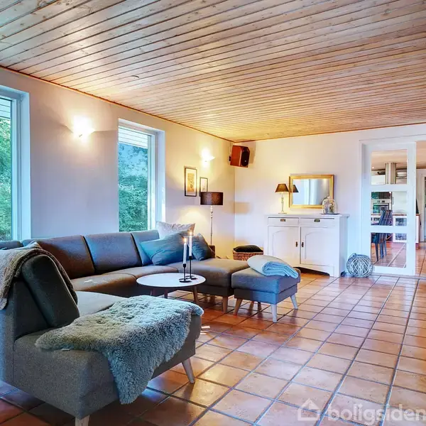 Stue med grå L-formet sofa står på et klinkegulv. Et lille bord med en lysestage er foran sofaen. Væggene er hvide med træloft, og store vinduer viser grønne omgivelser udenfor.