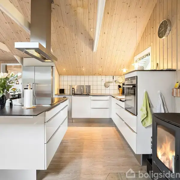 Køkken med hvide skabe og træloft, indeholder en brændeovn til højre og en stor emhætte til venstre. Planter og køkkenredskaber på bordet bidrager til hjemlig hygge.
