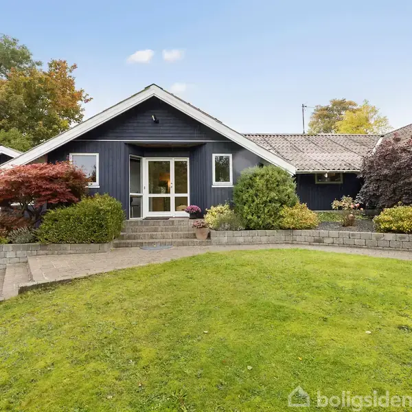 Mørkeblåt hus med hvide vinduesrammer står i en velplejet have med grøn græsplæne og farverige buske. Himmel er klar, omgivelserne fredfyldte.