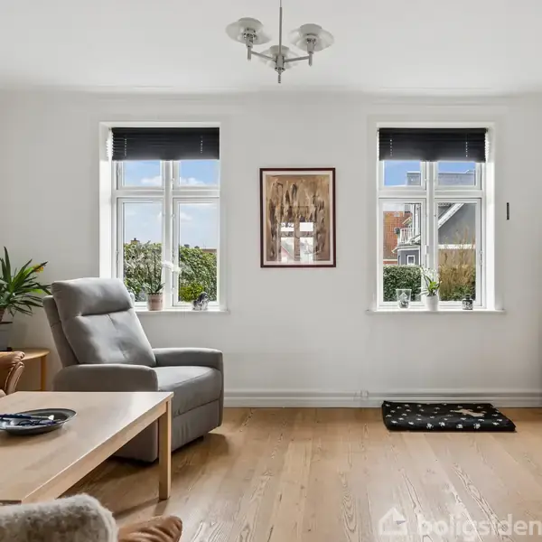 Stuen har en grå lænestol, et træbord og en lædersofa. To store vinduer giver udsigt til haven. Et TV står på et skab. Møblerne er moderne og rummet lyst.