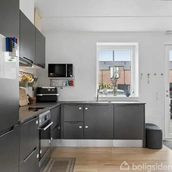 Et moderne rækkehus af mursten står stille. Foran huset er der en indkørsel og nogle grønne buske. I baggrunden ses en åben vej og græsarealer.