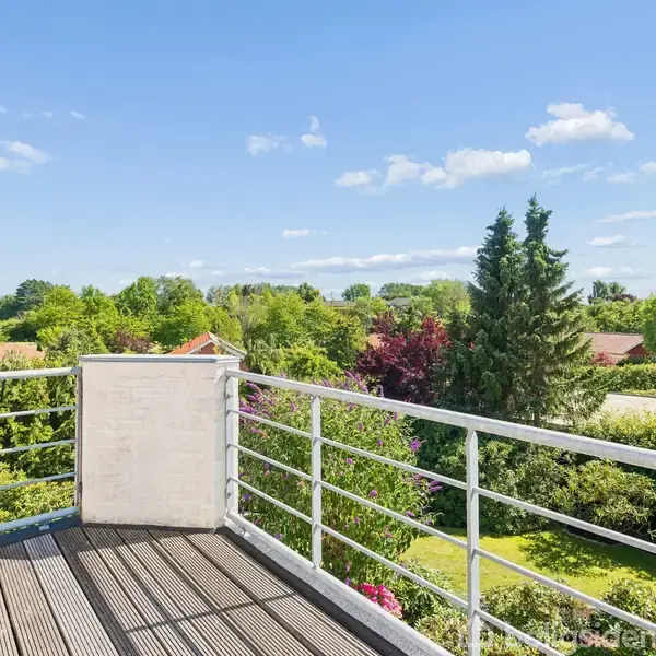En terrasse med træplanker og metalrækværk giver udsigt over en frodig have med forskellige træer og buske under en klar blå himmel.