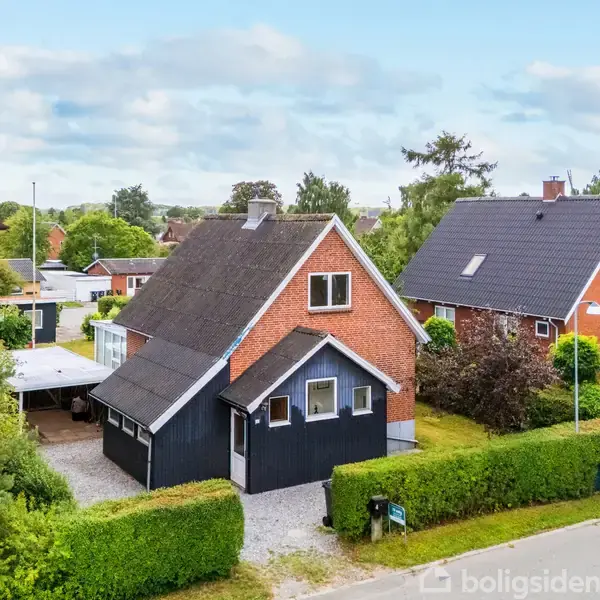 En rød murstensvilla med sort tag står på en velplejet ejendom omkranset af grønne hække. Et hvidt stakit og indkørsel ses foran huset, mens nabohusene ligger tæt omkring.