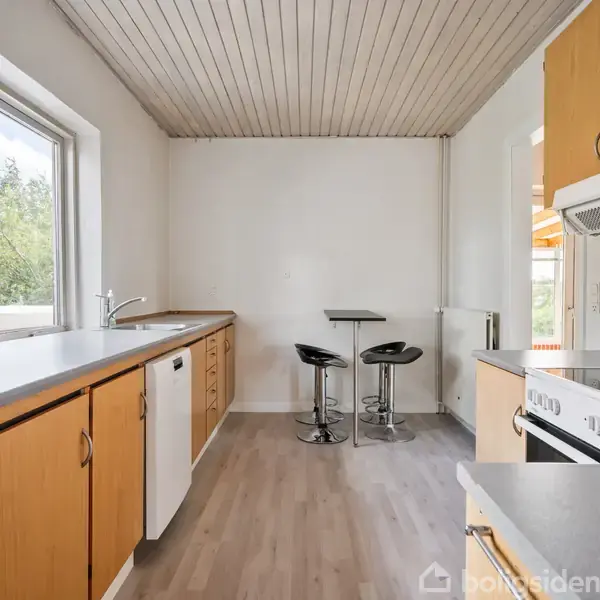 Et køkken med lyse trægulve og hvide vægge. Køkkenelementer i træ står langs væggen, med en ovn og emhætte til højre. To sorte barstole ved et lille bord.