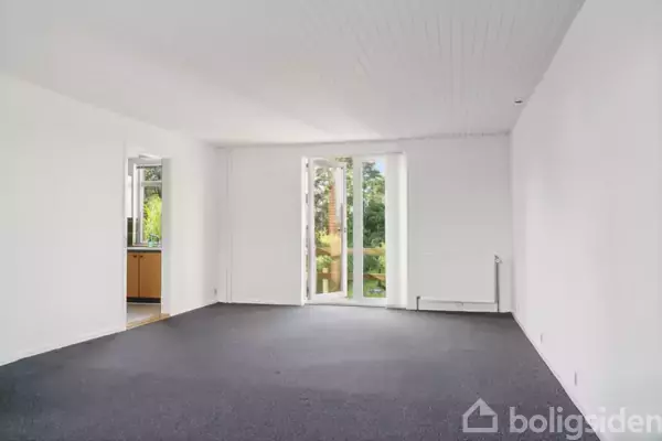 Et tomt rum med gråt gulvtæppe og hvide vægge; store vinduer åbner ud mod en grøn have. En dør forbinder rummet til et køkken.