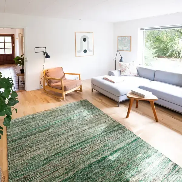 Stue med lysegrå sofa og træstol, står ved et stort vindue med udsigt til haven. Grøn tæppe på trægulv, omgivet af planter og lamper. Væggene er dekoreret med kunst.