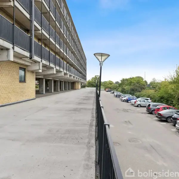 En lang, fleretagers bygning strækker sig langs venstre side. Foran er en bred promenade uden aktivitet. Til højre holder en række biler parkeret langs en grøn, træklædt kant.