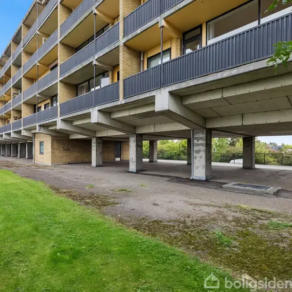 Lejlighedskompleks står stille, strukturen har åbne søjler i bunden, omgivet af græs og en parkeringsplads. Bygningen er flere etager høj med altaner på hver etage.