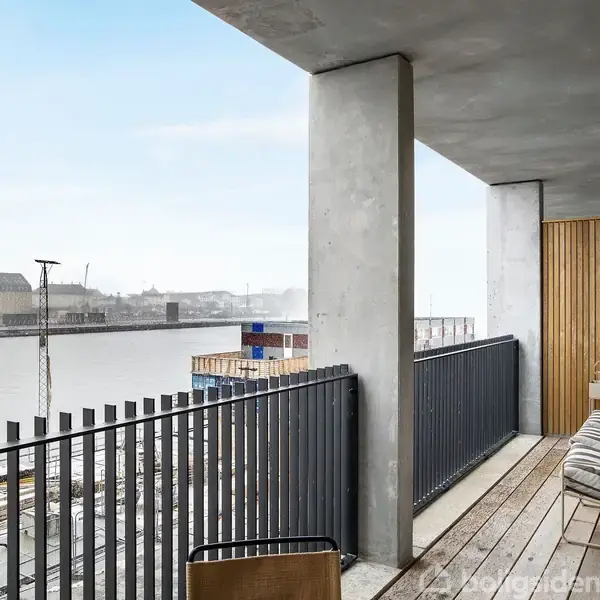 Altan med trægulv og to stole. Udsigt over en havn med vand, bygninger i baggrunden og grå himmel. Højt rækværk løber langs altanen. Ingen tekst til stede.