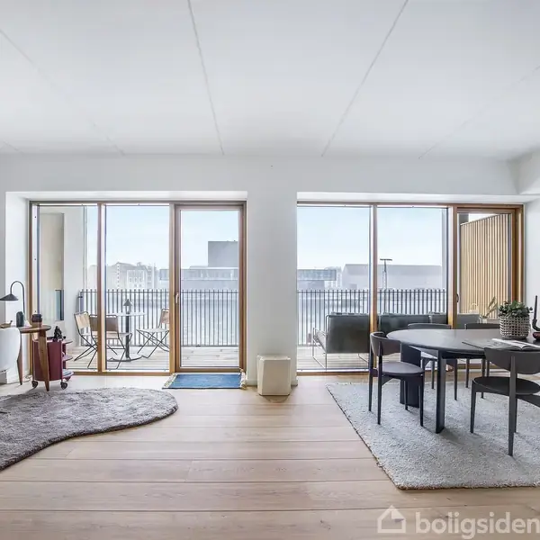 Stue med trægulv, en rund grå spisebord med stole til højre, og et skrivebord til venstre. Store vinduer i baggrunden giver udsigt til altan og bybillede.