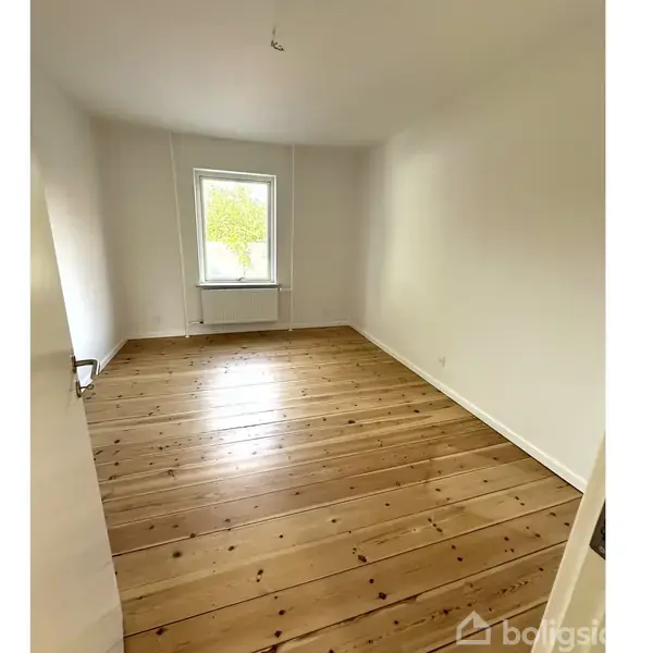 Et tomt værelse med lyst trægulv og hvide vægge. En radiator står under et enkelt vindue, som lukker dagslys ind fra det udendørs grønne område.