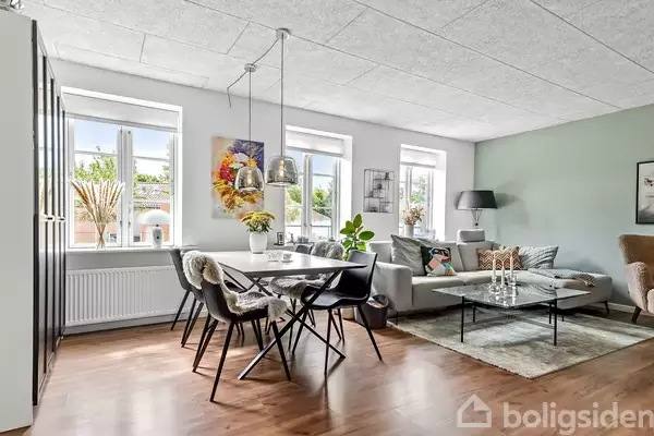 En stue med spisebord, der er dækket med stole og en vase med blomster. Sofa og sofabord står ved siden af med lamper omkring. Lys strømmer ind gennem flere vinduer.