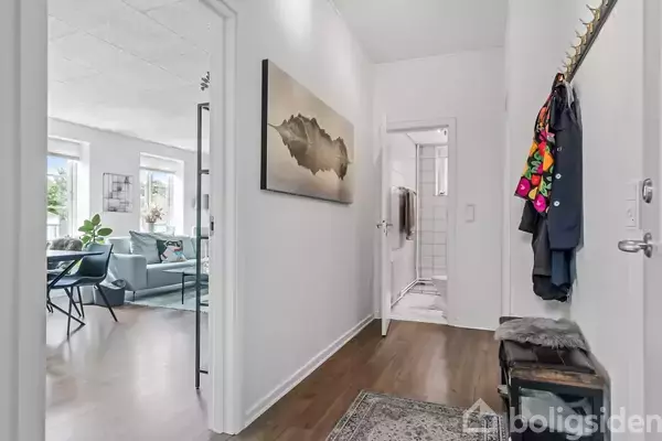 Gang med maleri på væggen, jakker hængende på knagerække, og udsigt til stue med blå sofa og spisestue. Trægulve og en dør fører til et badeværelse med hvide fliser.