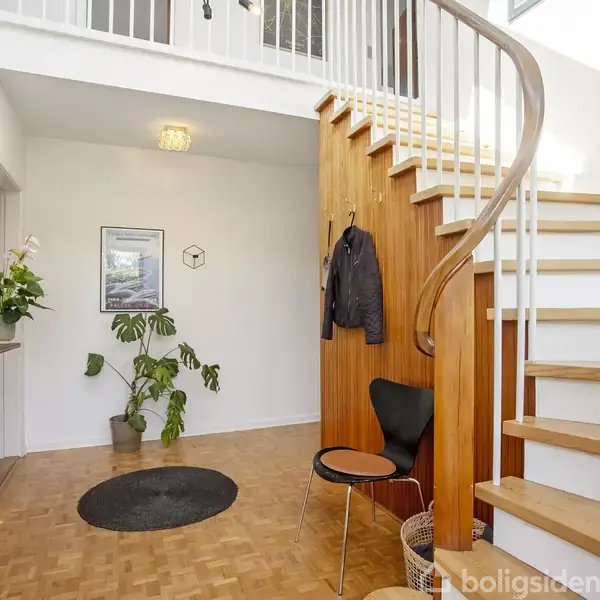 Entré med trætrappe til førstesalen, en stol og bord ved siden af jakke på væggen. Der er en rund vægplante og et maleri i baggrunden.