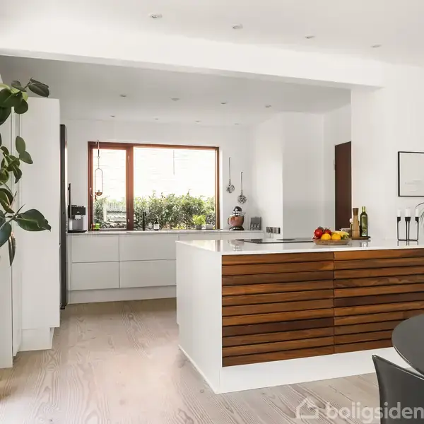 Køkken med træbeklædt køkkenø og hvide elementer, har planter og frugtskål. Stor vinduesudsigt bagved. Funktionelt og stilfuldt rum med spisebord til højre.