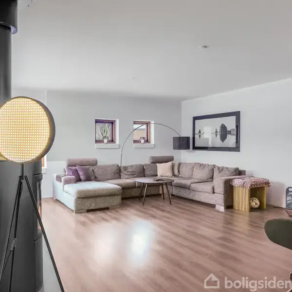 Gulvlampe lyser i stue, hvor en stor beige sofa står langs væggen. På væggen hænger to kunsttryk. En cykel ses ved siden af sofaen. Trægulv omkring.