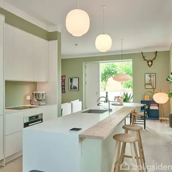 Køkken med hvidt, moderne interiør og en stor ø med barstole. Ved siden er der udsmykning med planter og vægkunst i en lys og indbydende atmosfære, og hjortegevir på væggen.