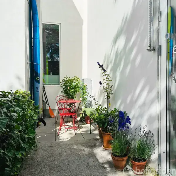 Rød metalstol og bord står i skyggefuld gårdhave omgivet af blomsterkrukker. Glasdør med teksten "69A" på højre side. Grønne planter vokser langs hvid væg. Paddleboard anes bagved.