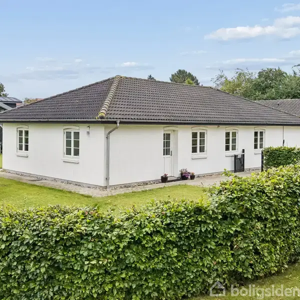 Hvid bungalow med mørkt tag står i rolig villakvarter omgivet af velplejet græsplæne og hække. Det er en énetages bygning med flere vinduer og grønt omkring.