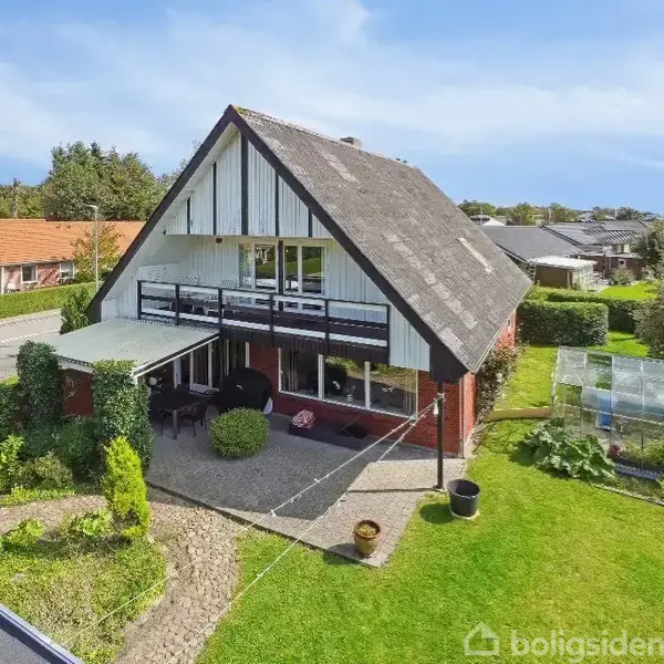 Et trekantet hus med store vinduer, omkranset af en velplejet have. Huset har en balkon og terrasse, mens drivhus og græsplæne ses i den solrige baghave.