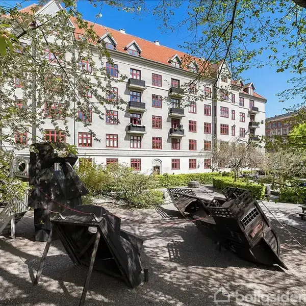 Stue med spisebord, stol og sofa, oplyst af lamper, i en hyggelig atmosfære. Billeder hænger på væggene, og et vindue åbner mod en balkon. En bogreol står i hjørnet.
