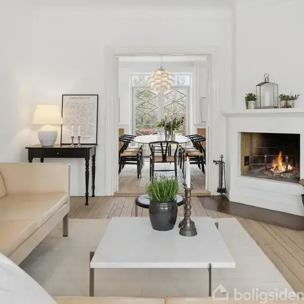 En stue med beige sofa, lavt sofabord med planter, brændende pejs til højre. I baggrunden ses et spisebord med stole og en lampe. Væggene er hvide.