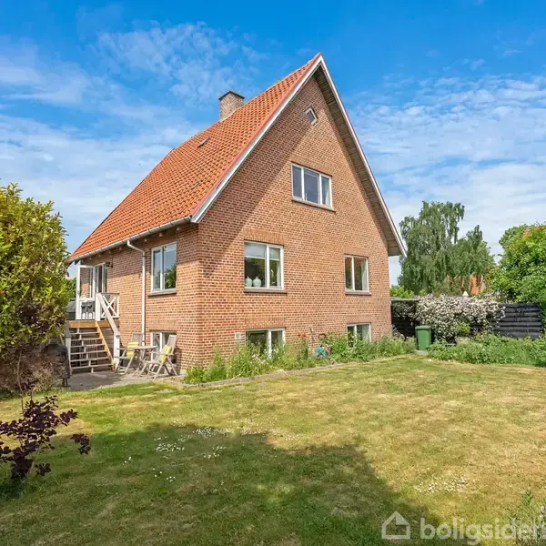 Et rødt murstenshus med saddeltag står i en solrig have. En hvid havebænk og et bord er placeret på græsplænen ved siden af huset.